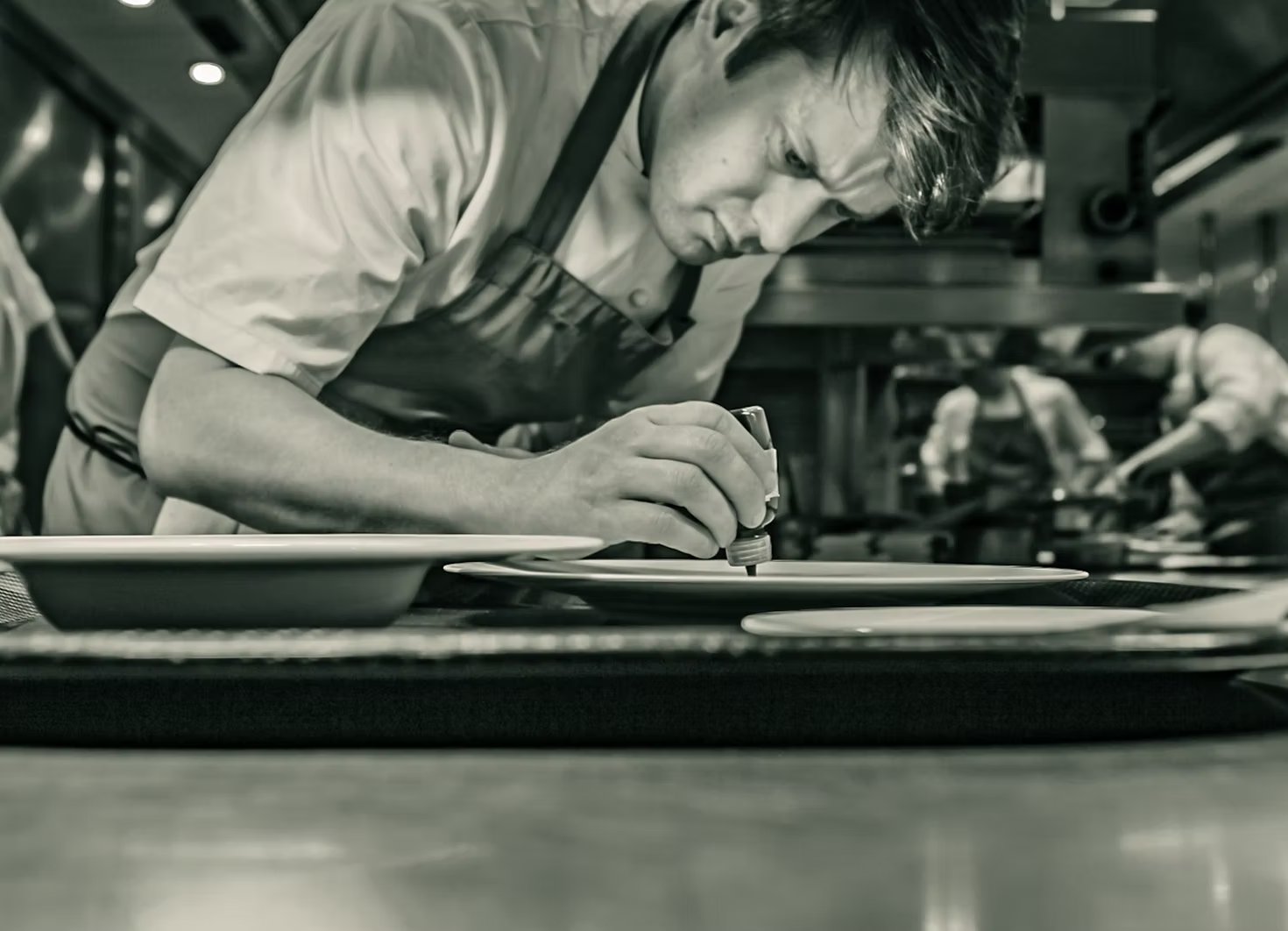 A chef plating with tweezers in a Michelin-starred kitchen, black and white